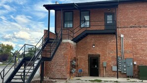 Un bâtiment en briques avec un escalier menant à un deuxième étage, une porte et des boîtiers électriques.