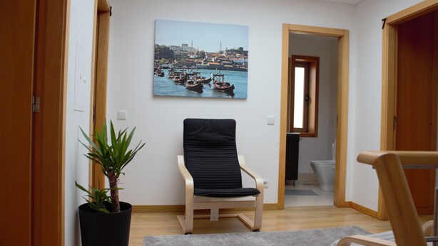 Una habitación con suelo de madera, una silla negra, una planta en maceta y un cuadro de una escena portuaria en la pared.