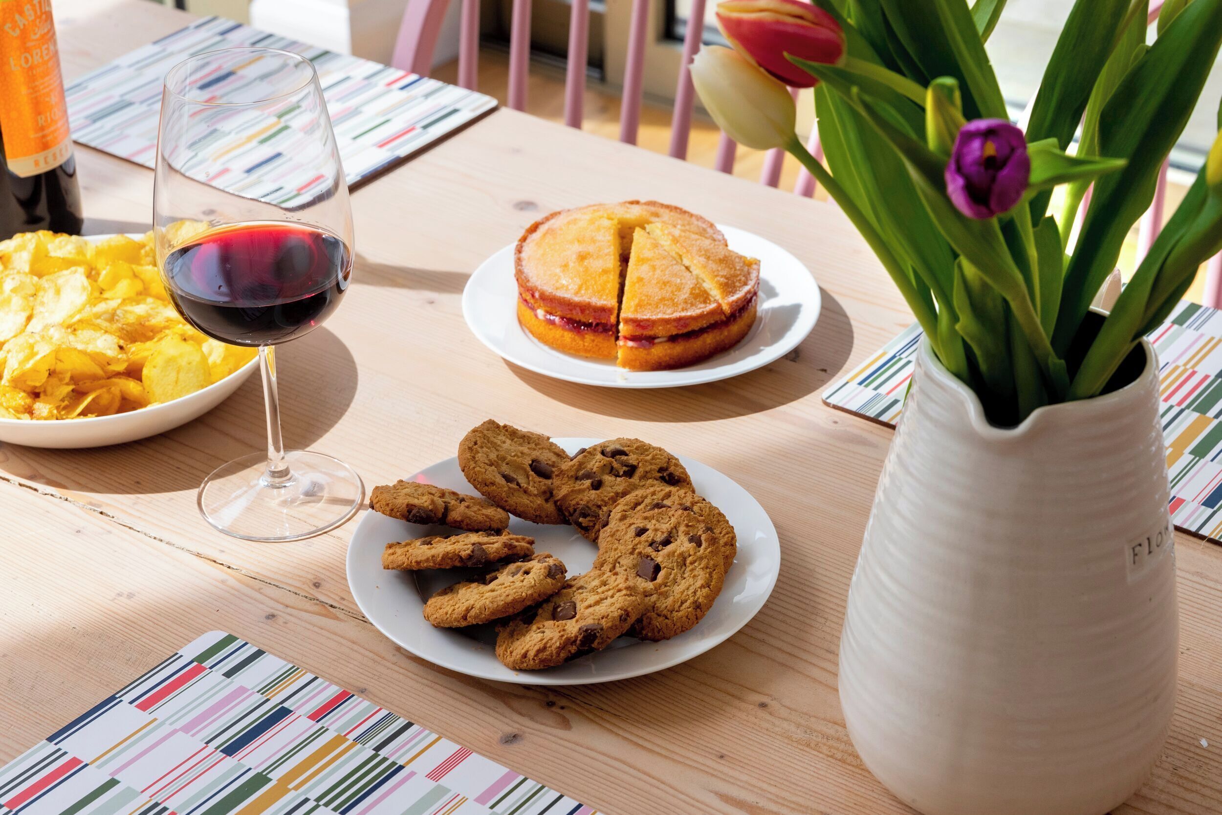 Ett bord med en tallrik kakor, ett glas rött vin, en tallrik med potatischips och en vas med blommor.