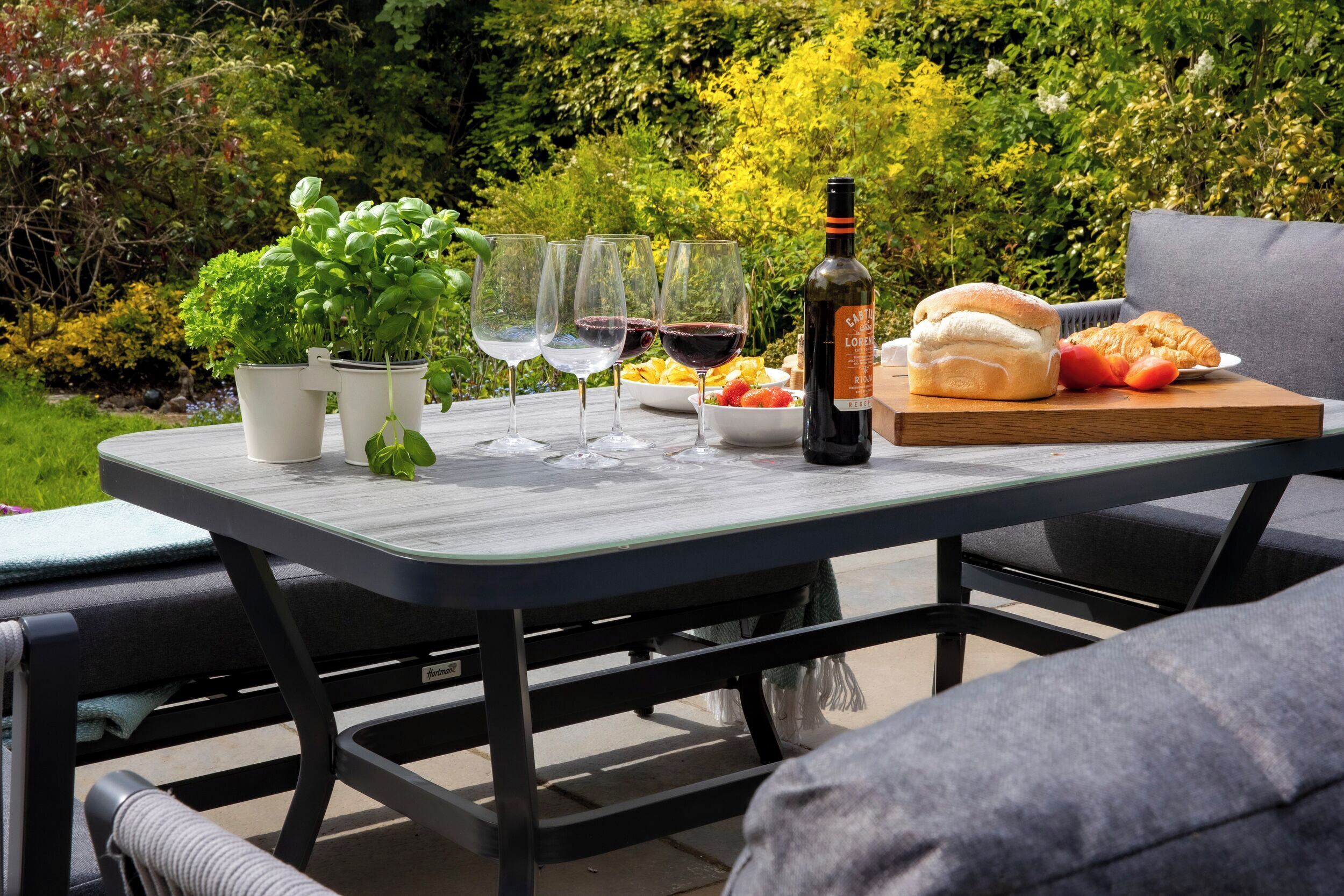 An outdoor dining table with a bottle, glasses, bread, and a bowl of fruit.