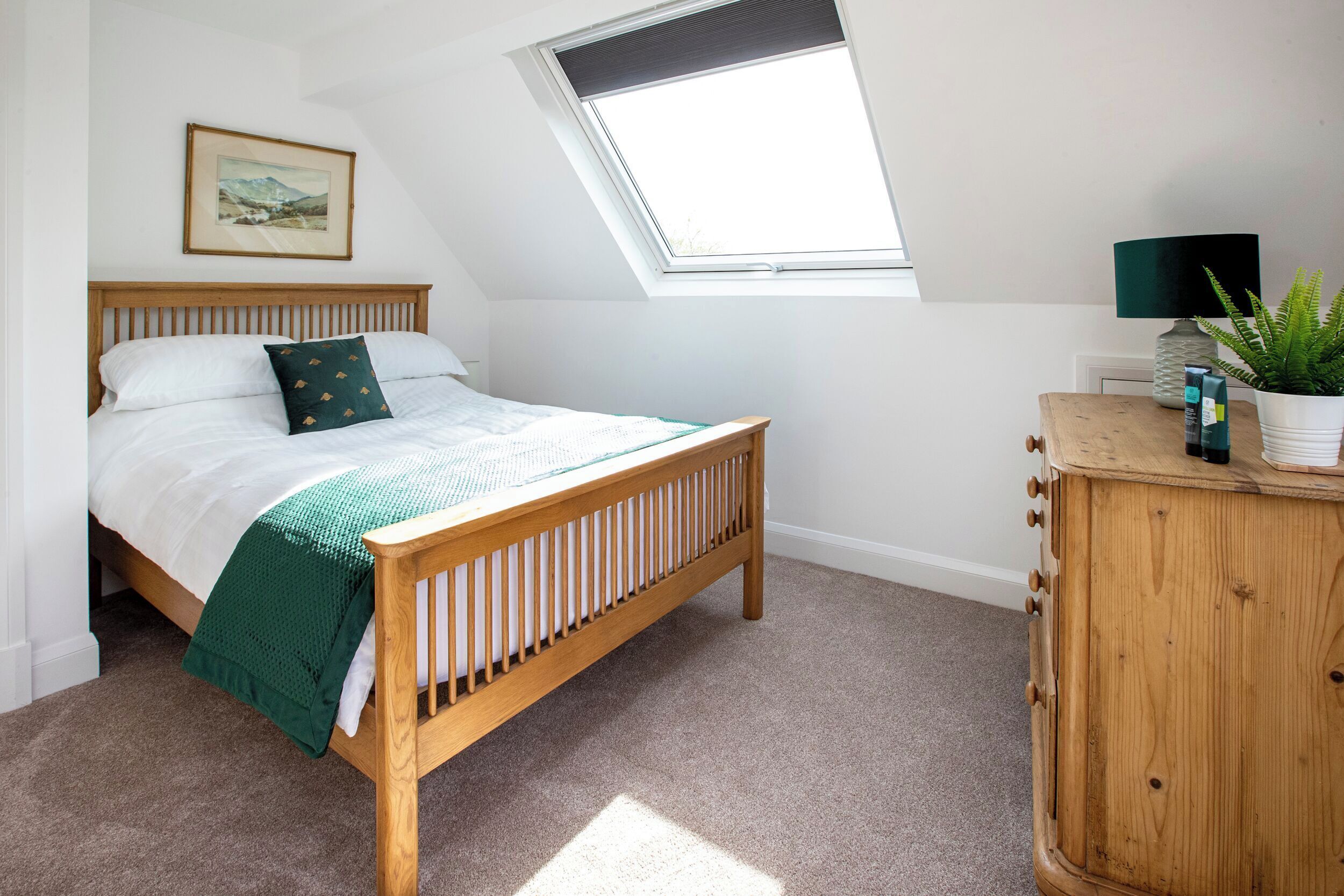 A bedroom with a wooden bed, a skylight, a bedside table with a lamp, and a framed picture on the wall.
