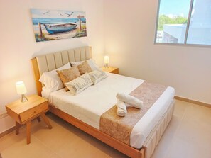 Un lit bien fait, avec une tête de lit, deux tables de chevet avec des lampes et un tableau représentant un bateau accroché au mur.