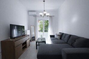 A modern living room with a flat-screen TV, a dark-colored sofa, a wooden cabinet, and a small table. - Piraeus Port Steps Away – Ground Floor Apt By ΜΡS (Pireas)