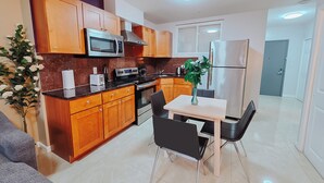 Een compacte keuken met houten kastjes, een magnetron, fornuis en een koelkast. Er staat een eettafel met stoelen en een vaas met een plantje.
