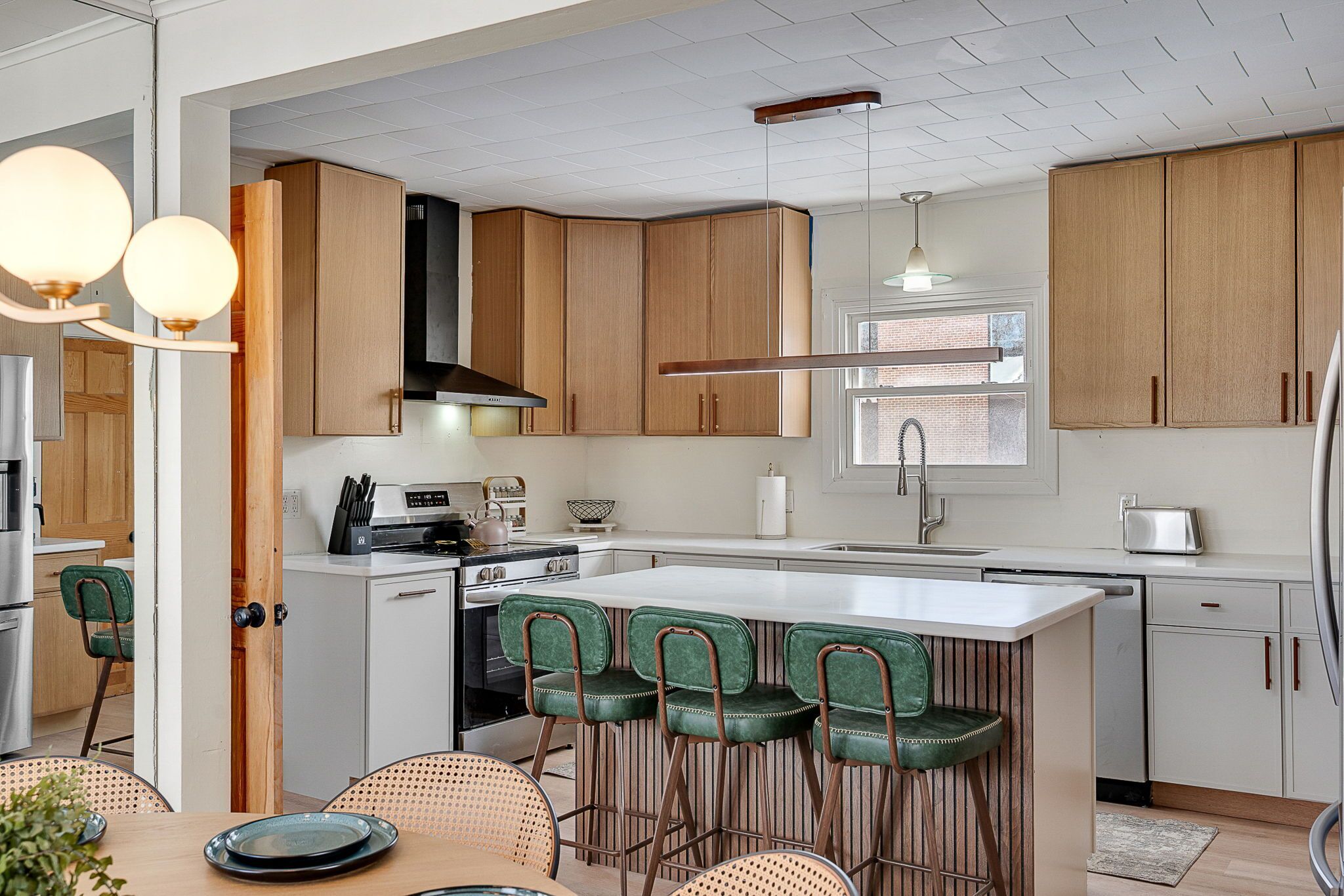 A modern kitchen with wooden cabinets, a central island with bar stools, a built-in oven, and a large window providing natural light.