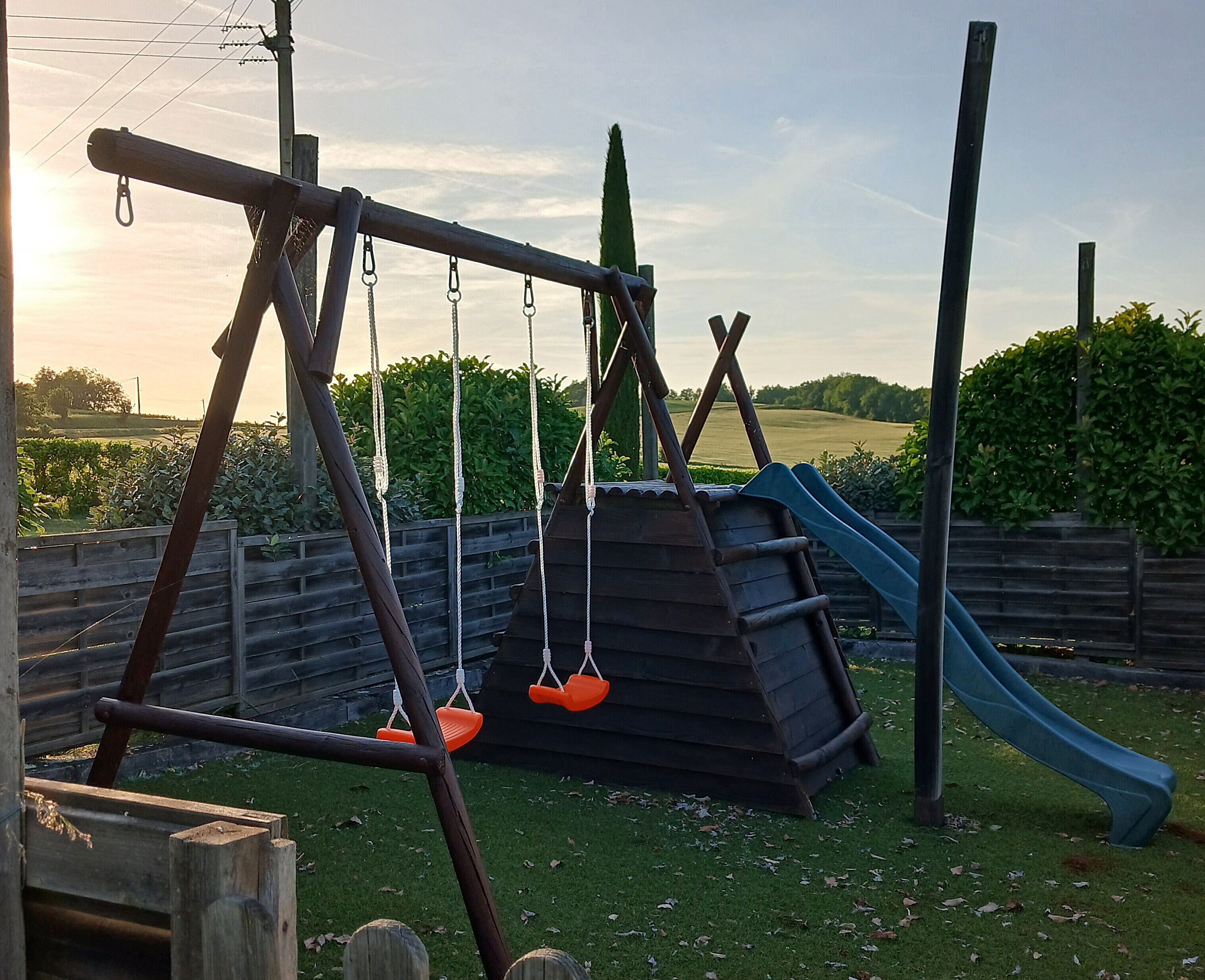 Ein Spielplatz mit einer Holzschaukel, orangenen Sitzen, einer Rutsche und einem umzäunten Bereich.