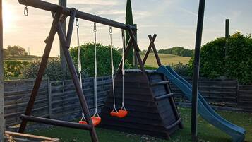 Ein Spielplatz mit einer Holzschaukel, orangenen Sitzen, einer Rutsche und einem umzäunten Bereich.