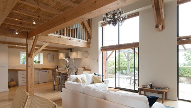 A spacious living area with a white sofa, wicker chairs, a wooden ceiling, and a large window offering a view of the outdoors.