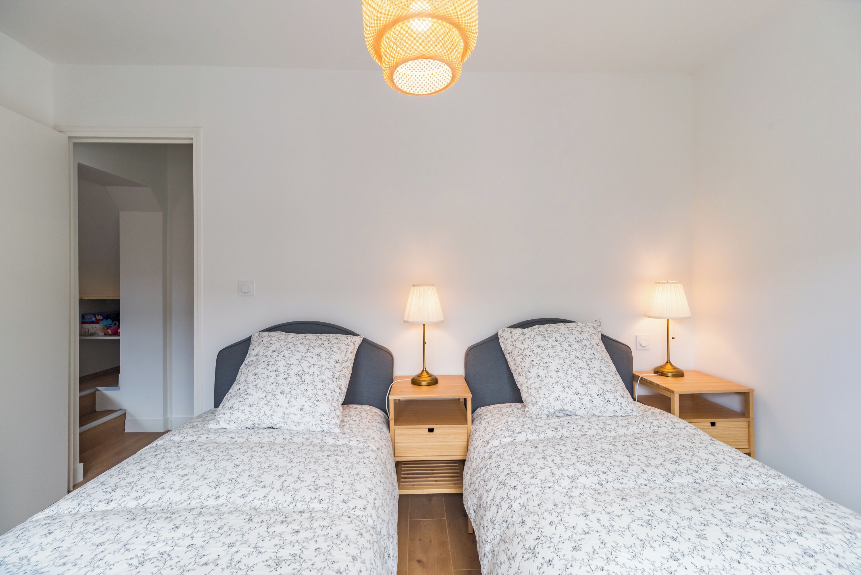 Une chambre avec deux lits, chacun étant doté d’une table de chevet et d’une lampe. On y trouve un placard dont la porte est ouverte sur un escalier.