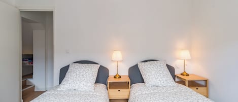 Une chambre avec deux lits, chacun étant doté d’une table de chevet et d’une lampe. On y trouve un placard dont la porte est ouverte sur un escalier.