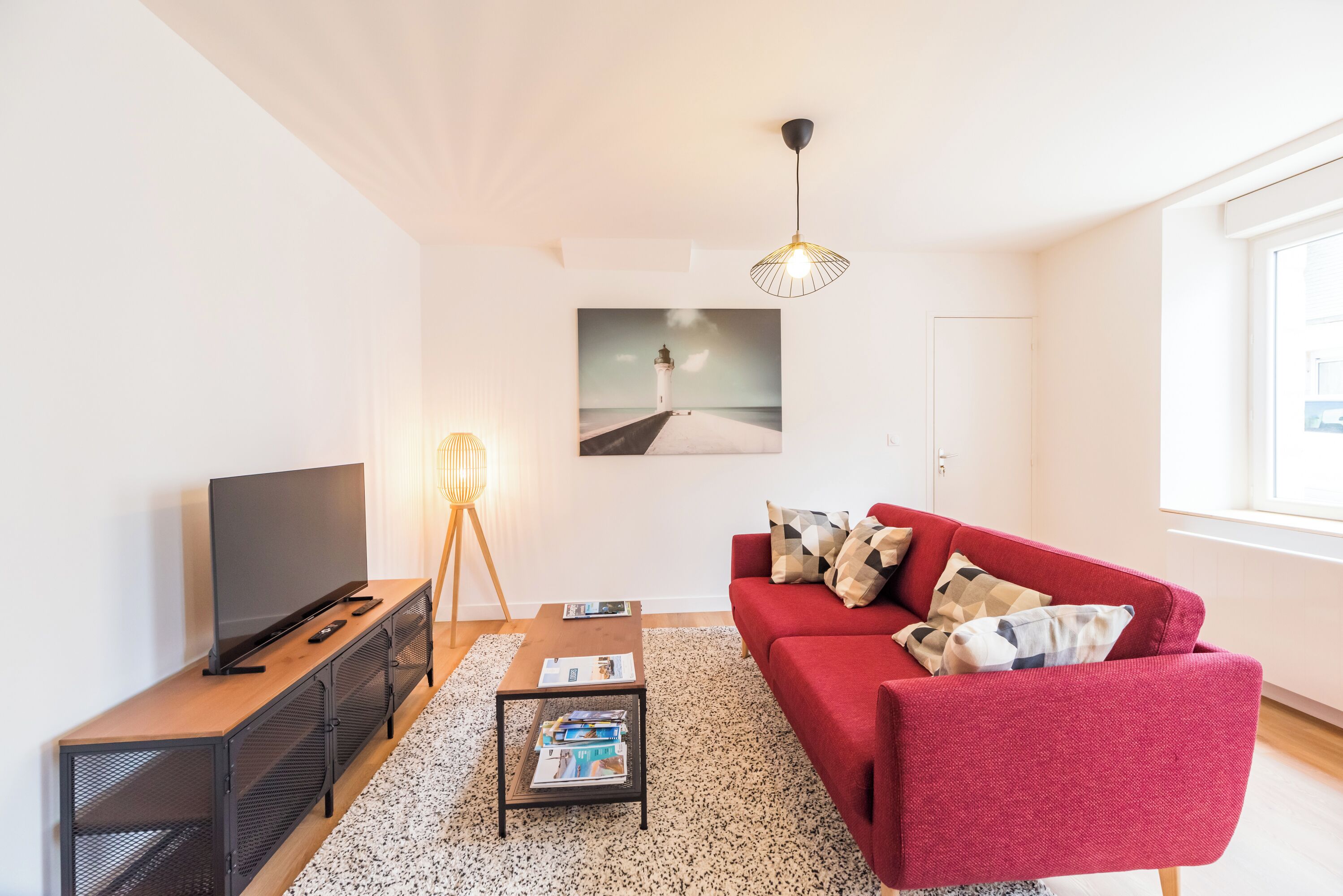 A modern living room with a red sofa, a TV on a wooden console, a standing lamp, and a picture of a lighthouse on the wall.