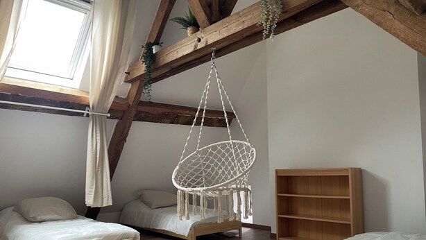 Een slaapkamer met twee houten bedden, een macramé hangstoel, een houten plank en een raam met gordijnen.