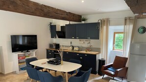 Een kleine keuken met donkere kastjes, een wit aanrechtblad en een ingebouwde televisie. Er is een eettafel met blauwe stoelen en een bijzettafeltje.