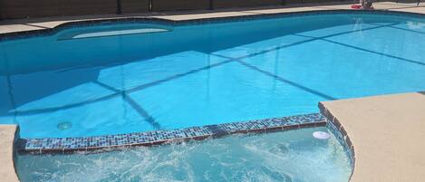 En swimmingpool med flisecant, omgivet af et hegn, og en basketballkurv i baggrunden.