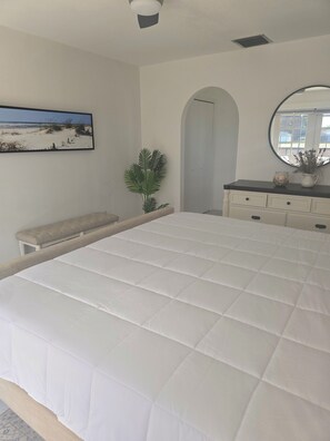 Ein Schlafzimmer mit einem großen Bett, einer Bank, einem Blumentopf, einem Schminktisch, einem Spiegel und einem gerahmten Bild an der Wand.