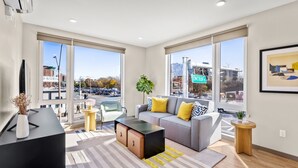 A modern living room with a sofa, coffee table, and a TV stand. Large windows offer a view of the street. - Elysian Residences Boston (Boston)
