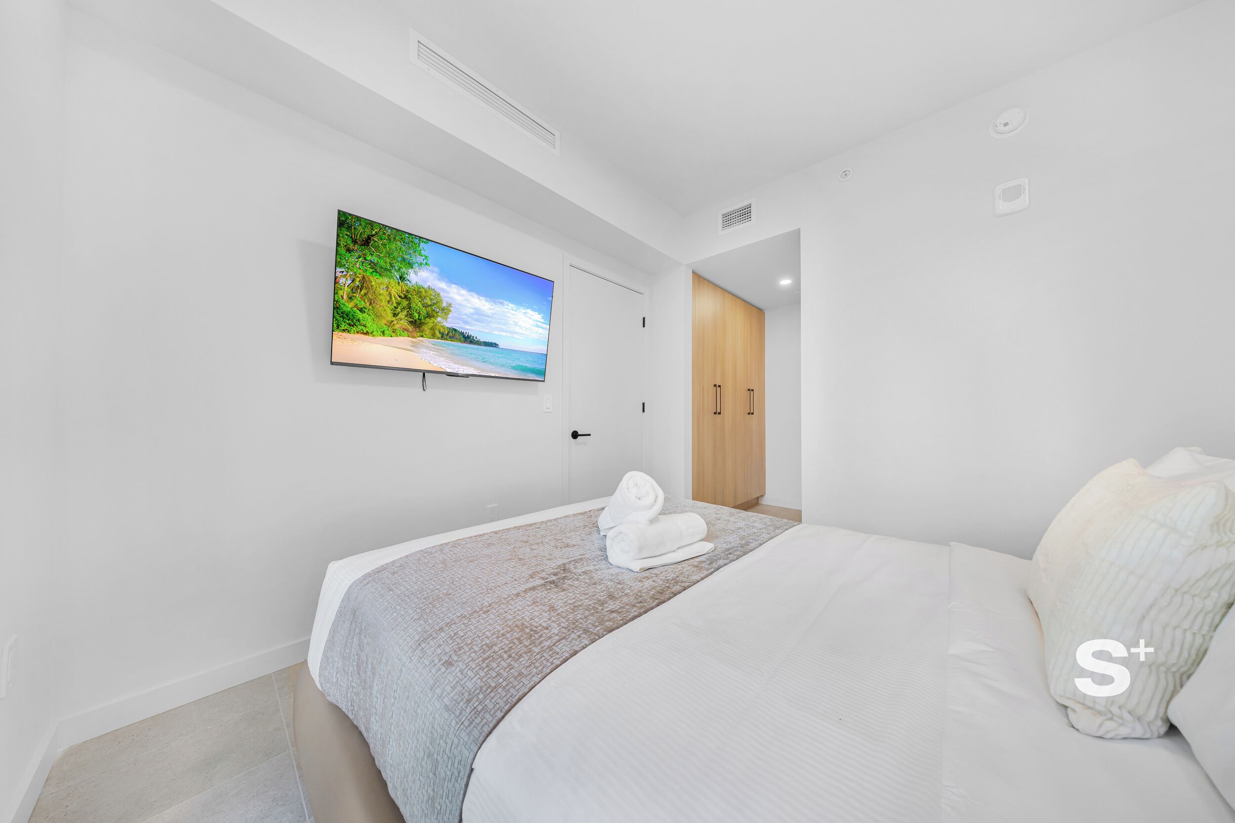 A modern bedroom with a large bed, a flat-screen TV mounted on the wall, and a wooden wardrobe.