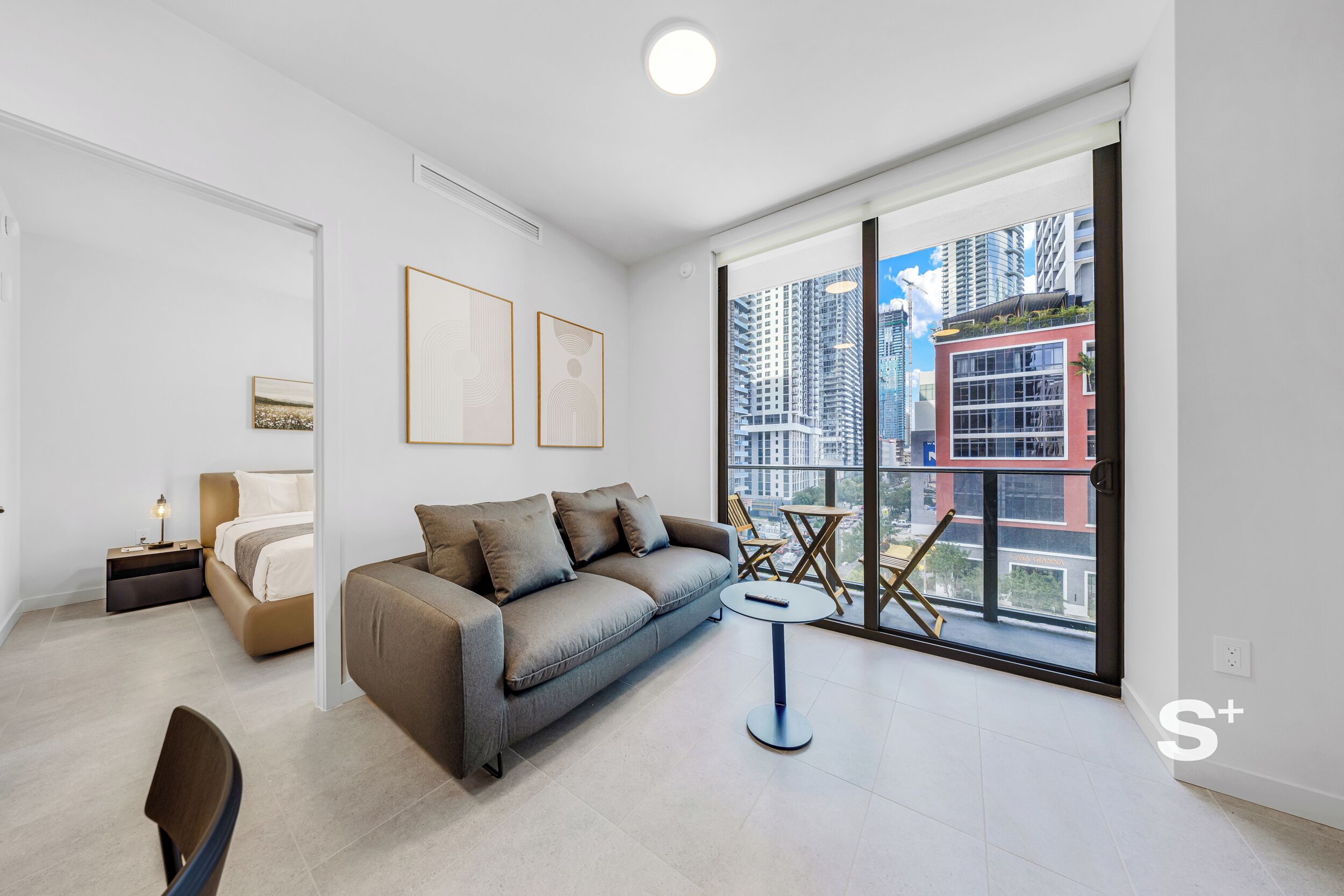 A modern living room with a sofa, a small round table, and a large window offering a city view.