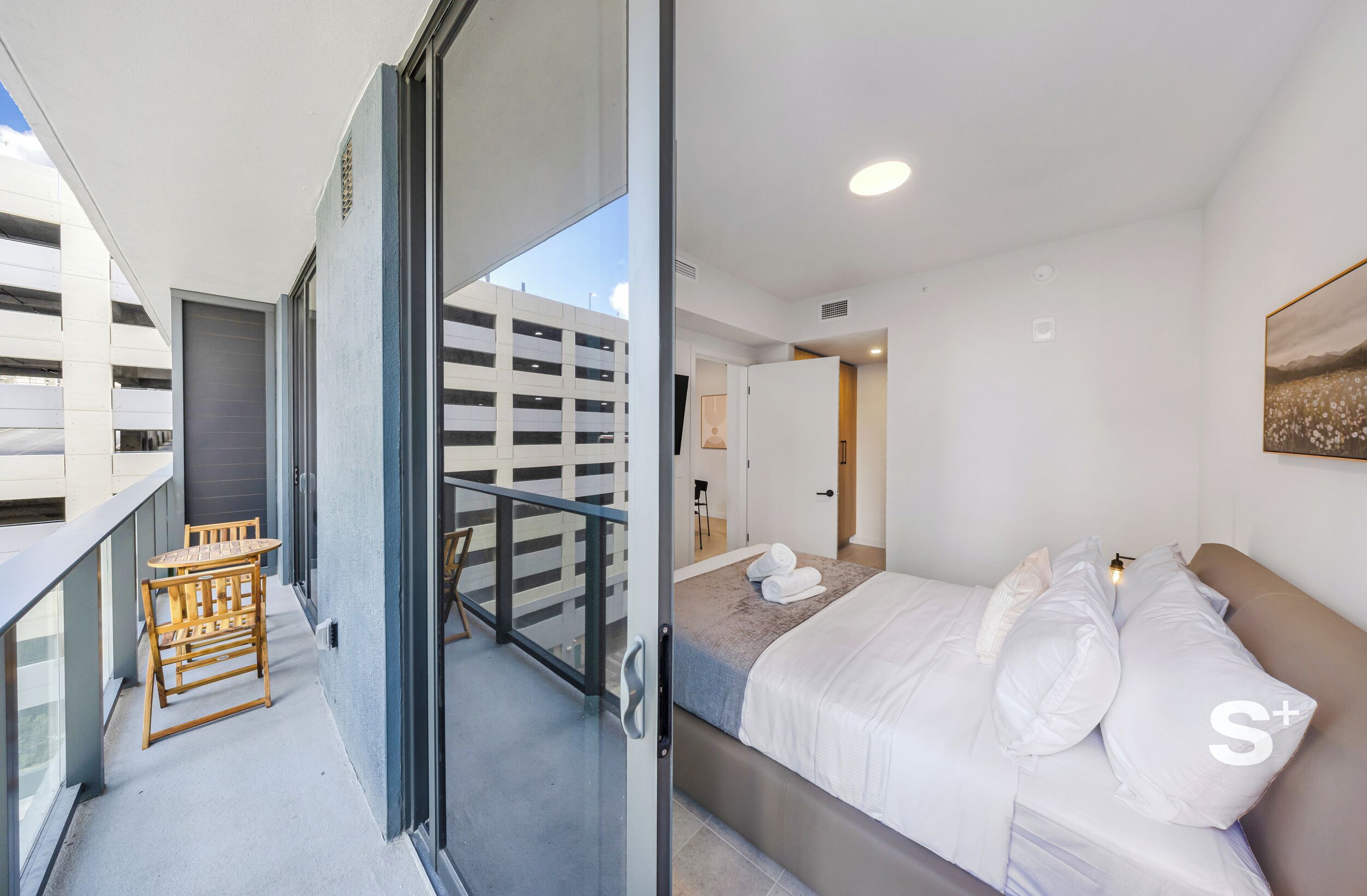 Ein modernes Hotelzimmer mit Balkon, einem Bett mit weißer Bettwäsche und Blick auf eine Parkgarage.