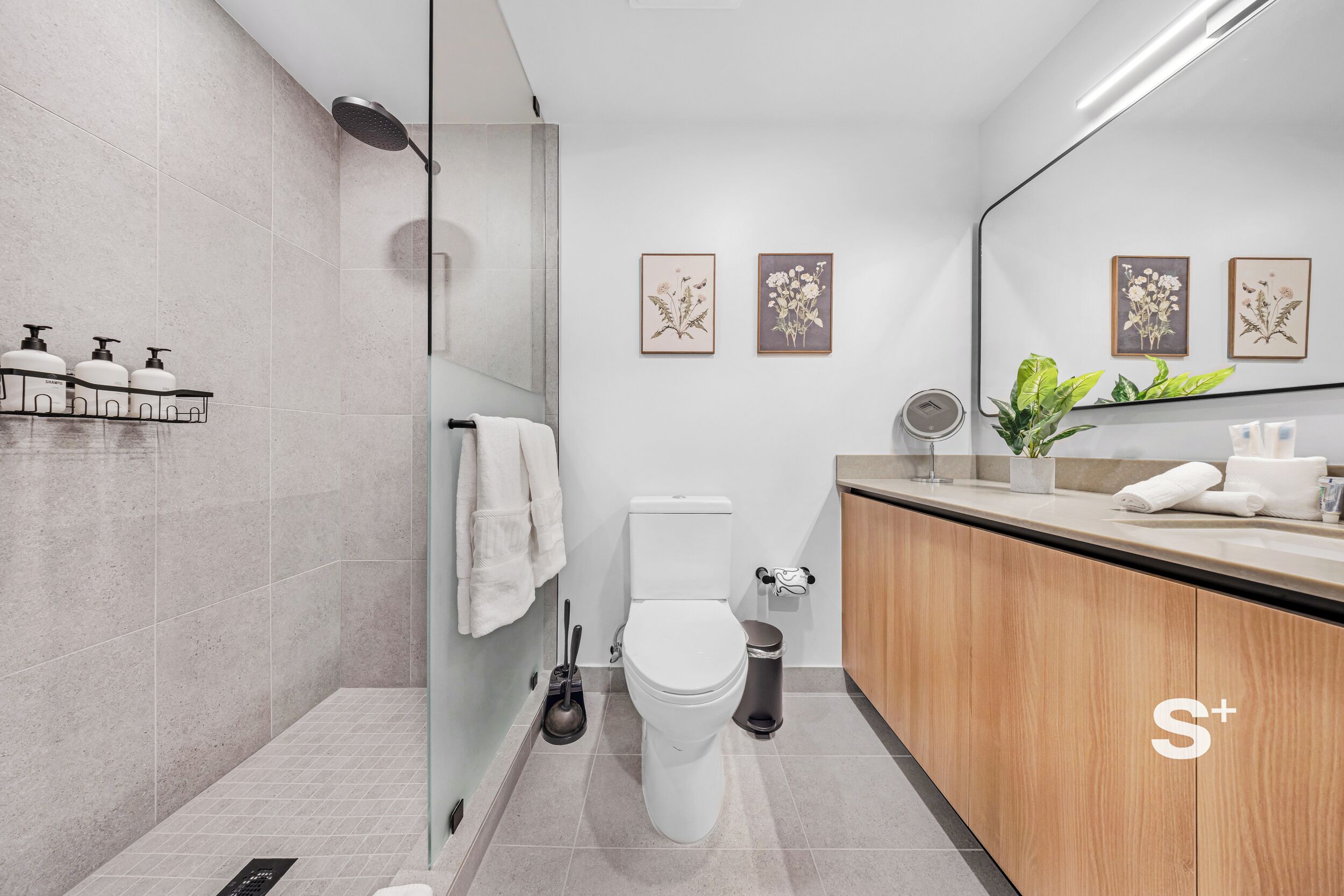 A modern bathroom with a toilet, bidet, and shower. There are framed botanical prints on the wall, a sink with a mirror, and a plant on the counter.