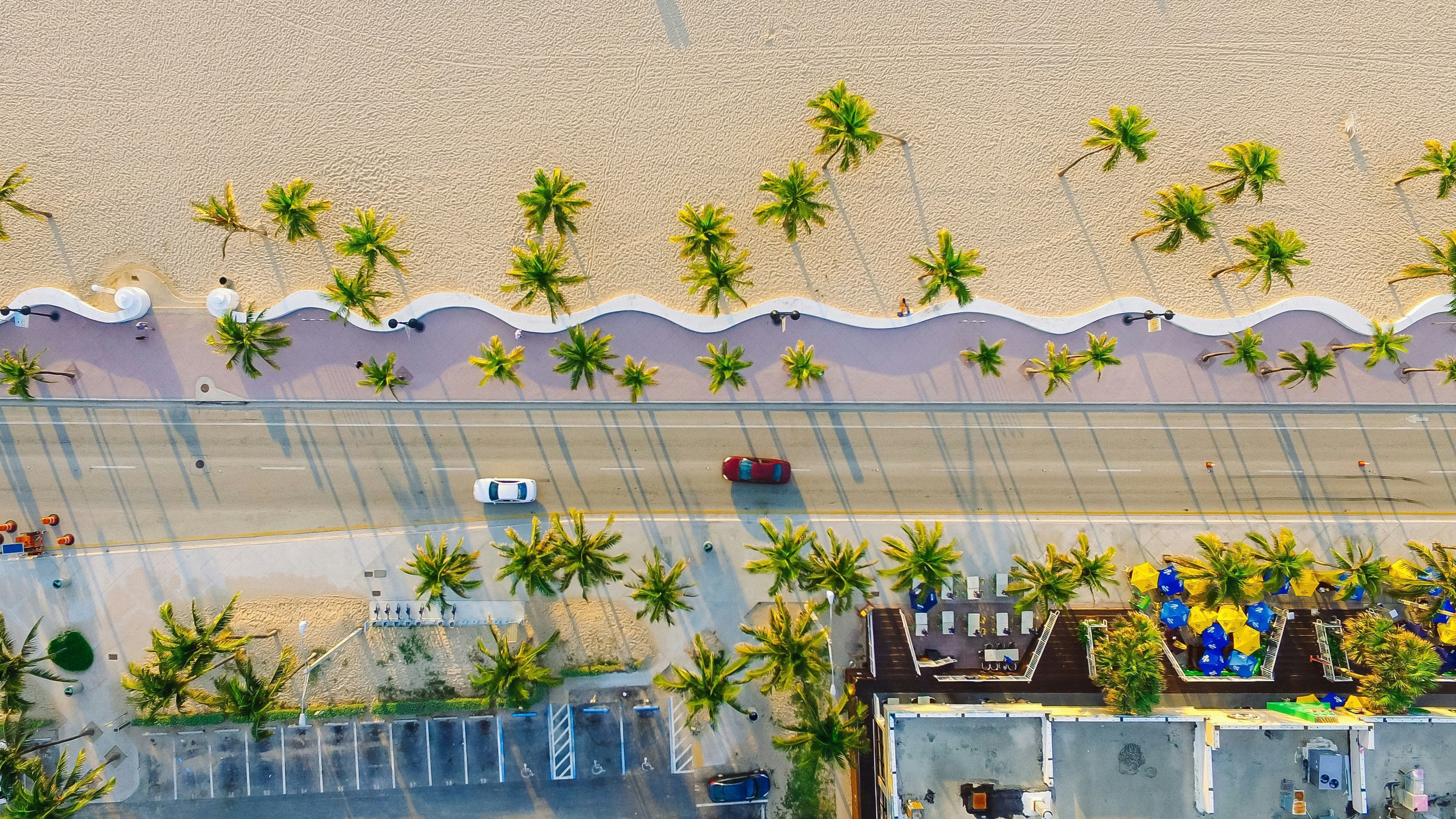 Luftaufnahme einer Strandgegend mit einer Straße, Parkplätzen und einem Gebäude mit Außenbereich.