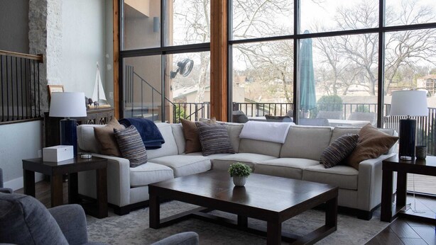 A living room with a sectional sofa, a coffee table, and a large window with a view of trees and buildings. - Spacious and fully loaded 4 Bed 4 Bath with Amazing Views on Grand Lake. (Afton)