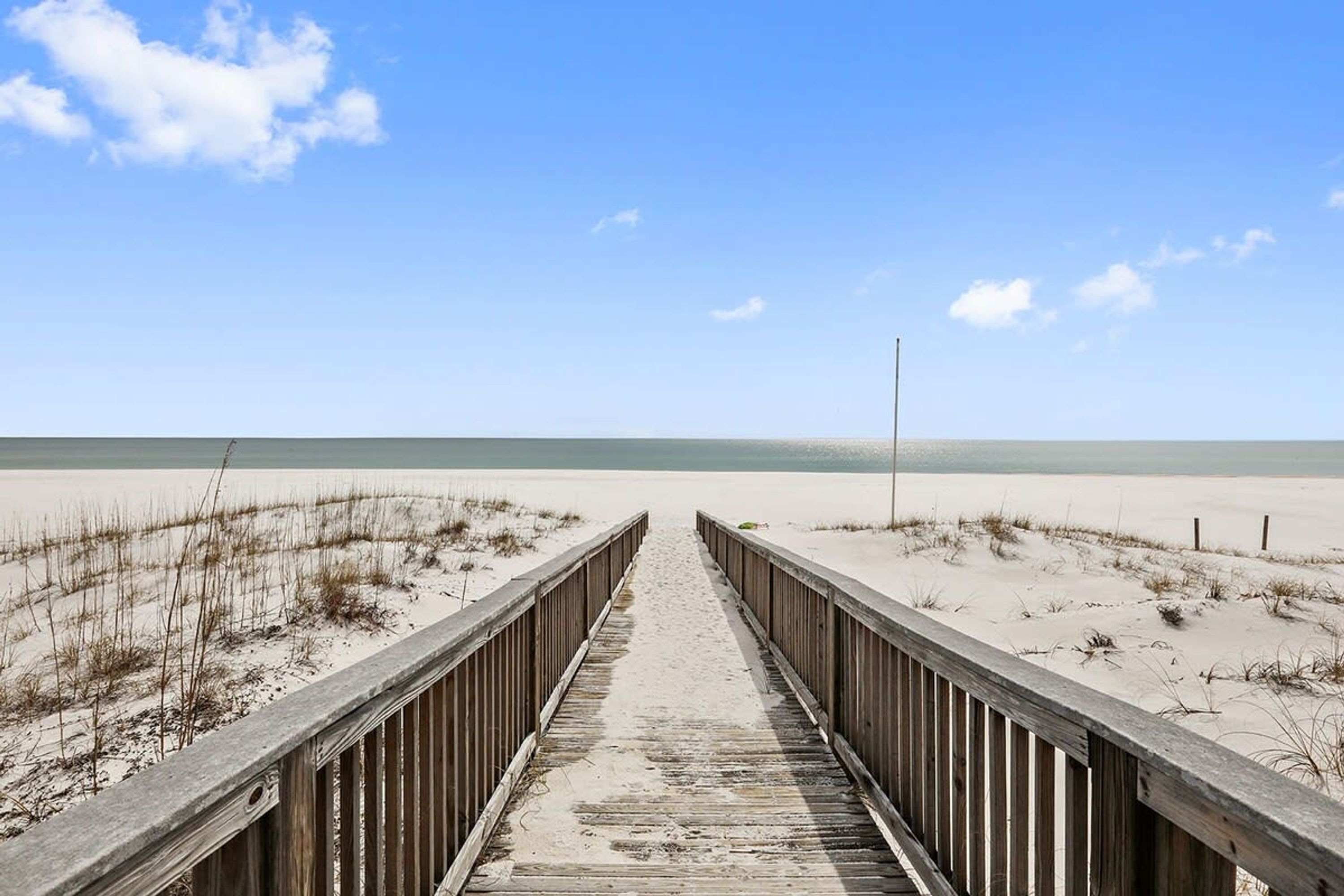 A wooden boardwalk leading to a beach with sand dunes and a clear blue sky.