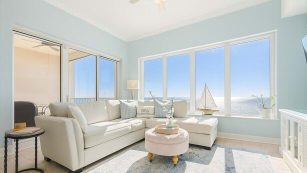 A living room with a light blue wall, a white sofa, a round ottoman, and a view of the ocean through large windows. - Regency Isle 1104 Penthouse With 3 Bedrooms (Orange Beach)