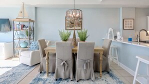 A dining area with a wooden table, four chairs, a chandelier, and a bookshelf. - Regency Isle 1104 Penthouse With 3 Bedrooms (Orange Beach)
