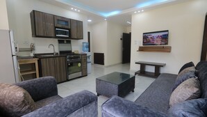 A modern living room with grey sofas and a glass coffee table. A kitchen with dark wood cabinets and a built-in microwave is visible in the background. - 7th Circle Diamnod (Amman)