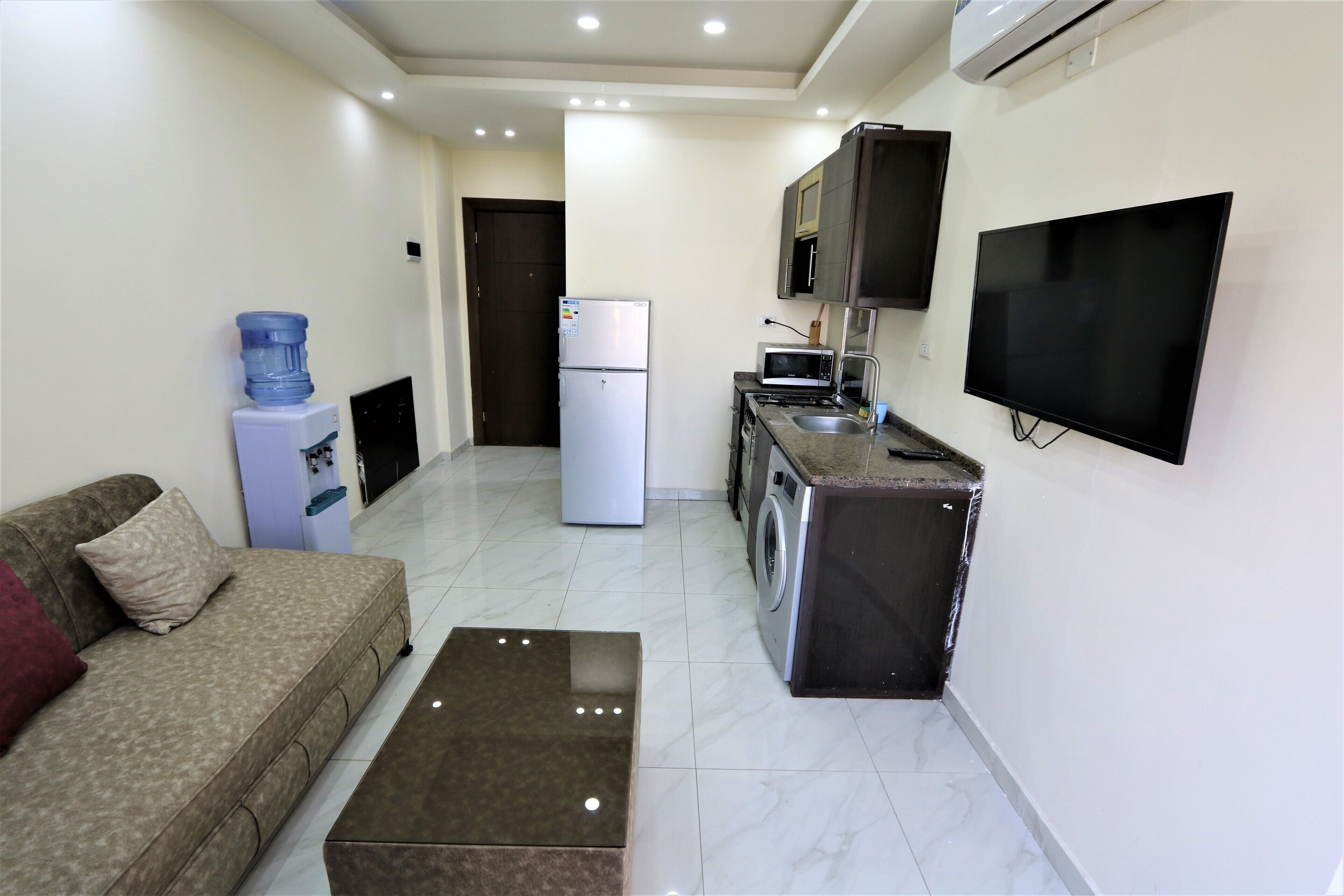 A compact living space with a sofa, a coffee table, a refrigerator, a microwave, a washing machine, and a TV mounted on the wall.