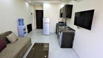 A compact living space with a sofa, a coffee table, a refrigerator, a microwave, a washing machine, and a TV mounted on the wall.