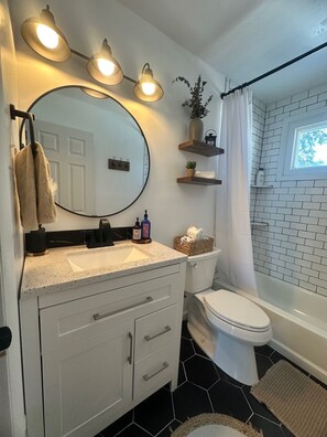 Une salle de bain avec un lavabo, des toilettes et une baignoire, tous de couleur blanche. Trois appliques murales sont fixées au-dessus du lavabo, ainsi qu’un grand miroir et une plante en pot, placée sur une étagère.