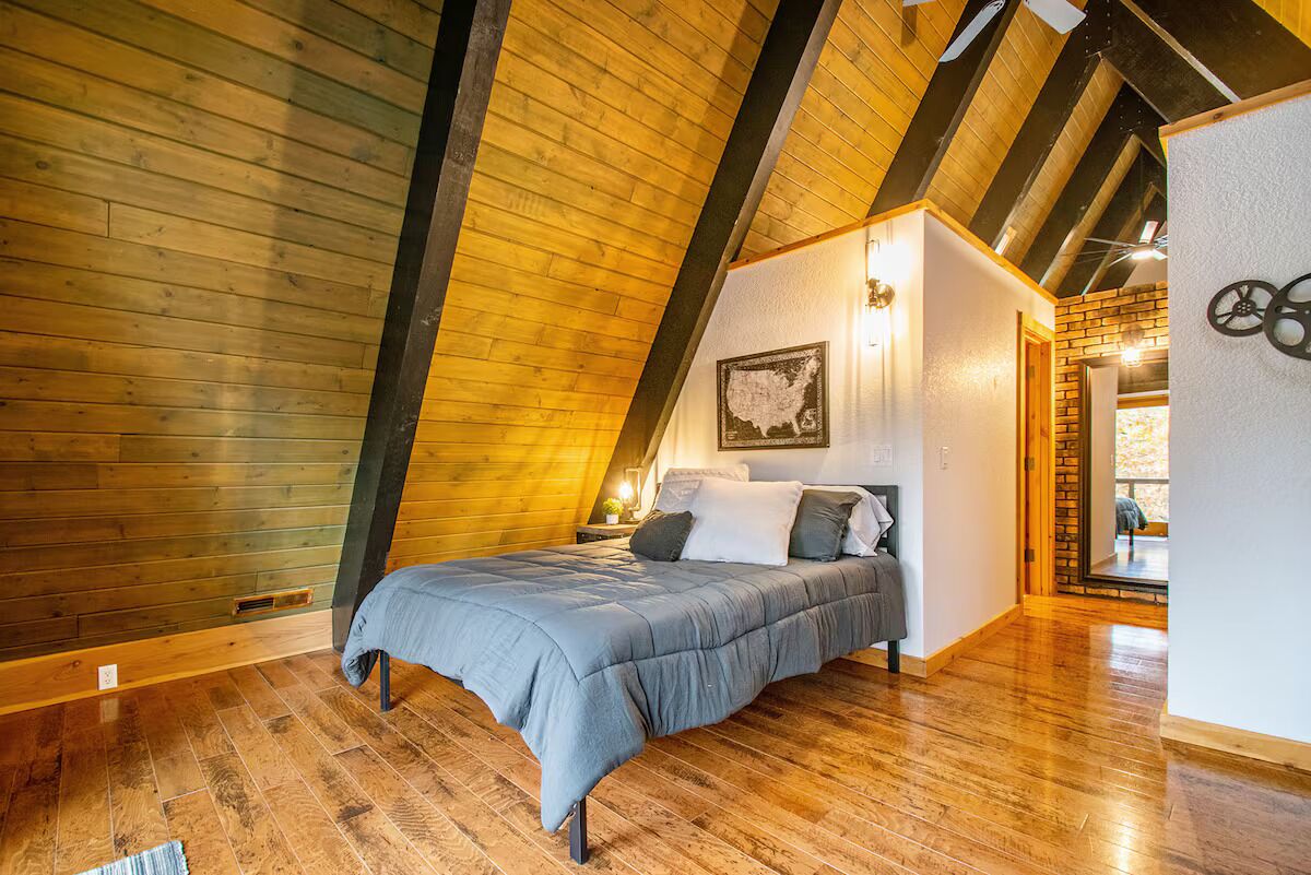 Une chambre avec un plafond en pente, un plancher en bois, un lit avec une couette grise et une carte accrochée au mur.
