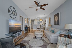 A modern living room with a flat-screen TV, a ceiling fan, and a dining area with a kitchen in the background. - Sunset Beach Escape! (Sunset Beach)