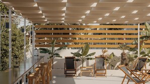 An outdoor seating area with wooden chairs and tables, a pergola overhead, and a view of a palm tree. - Cavo Notos Beach Hotel (Lefkimmi)