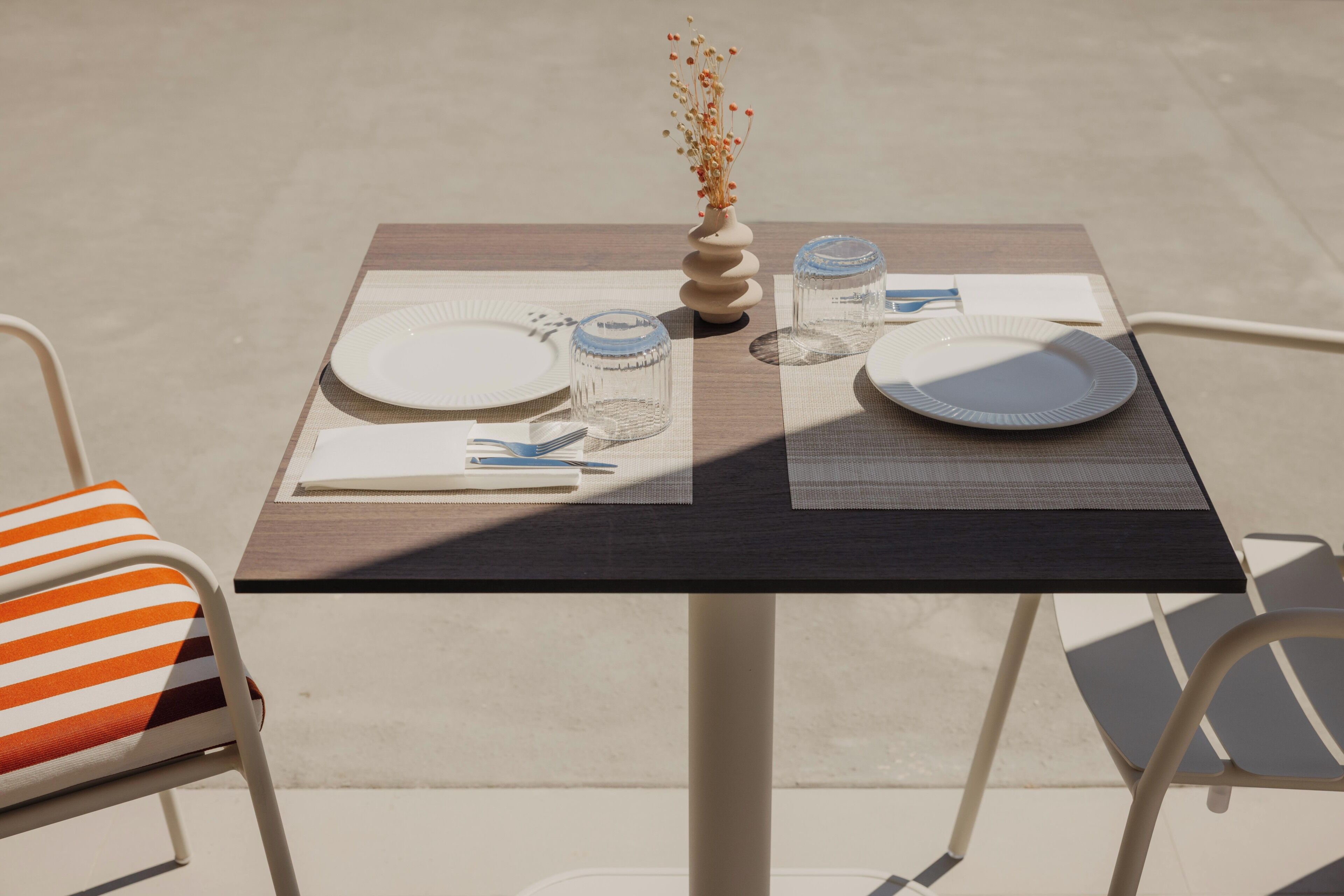 A table set for two with plates, cutlery, and a vase with dried flowers.