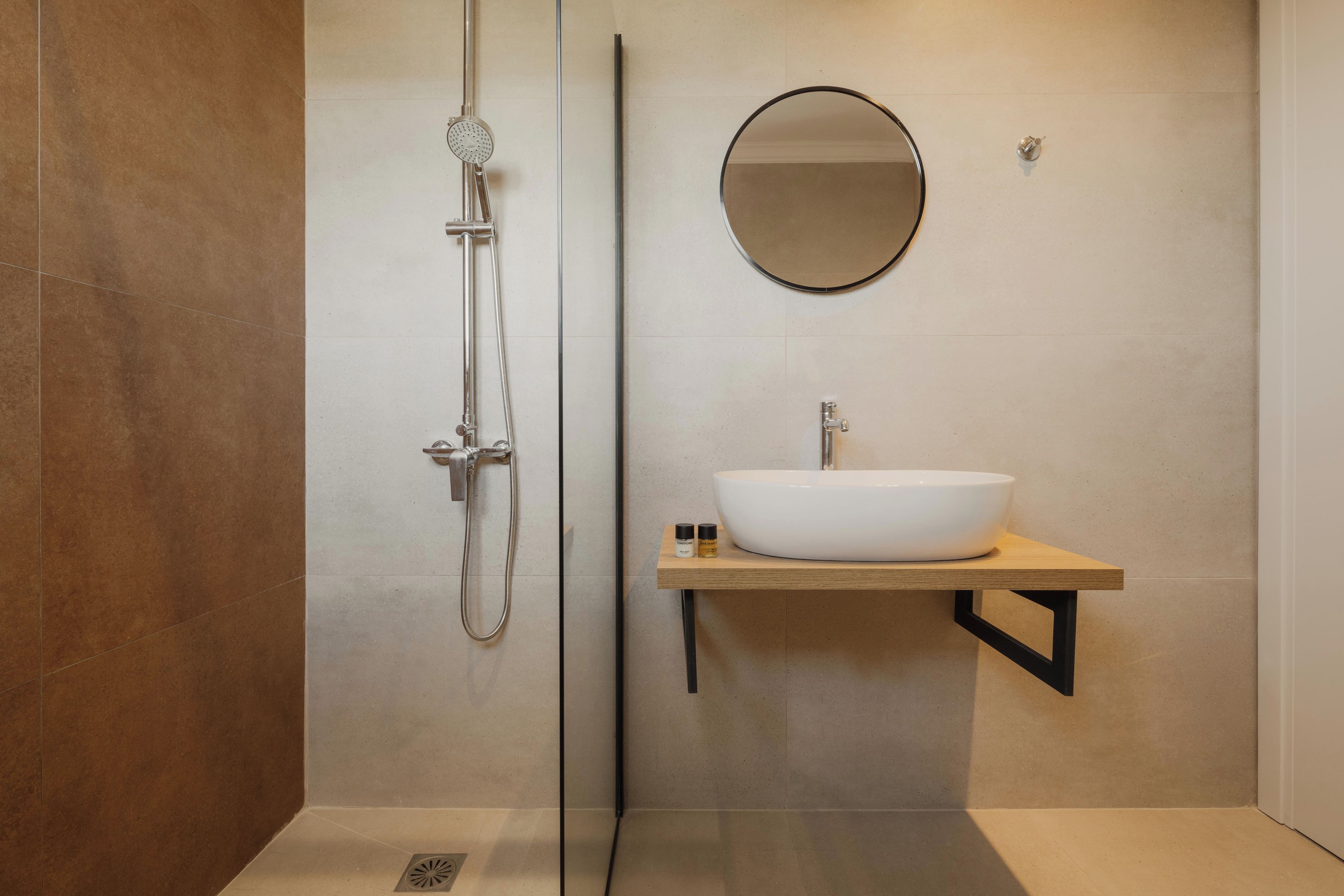 A modern bathroom with a glass-enclosed shower, a round mirror, and a white sink on a wooden shelf.
