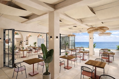 A spacious outdoor dining area with multiple wooden tables and chairs, large potted plants, and a view of the ocean.
