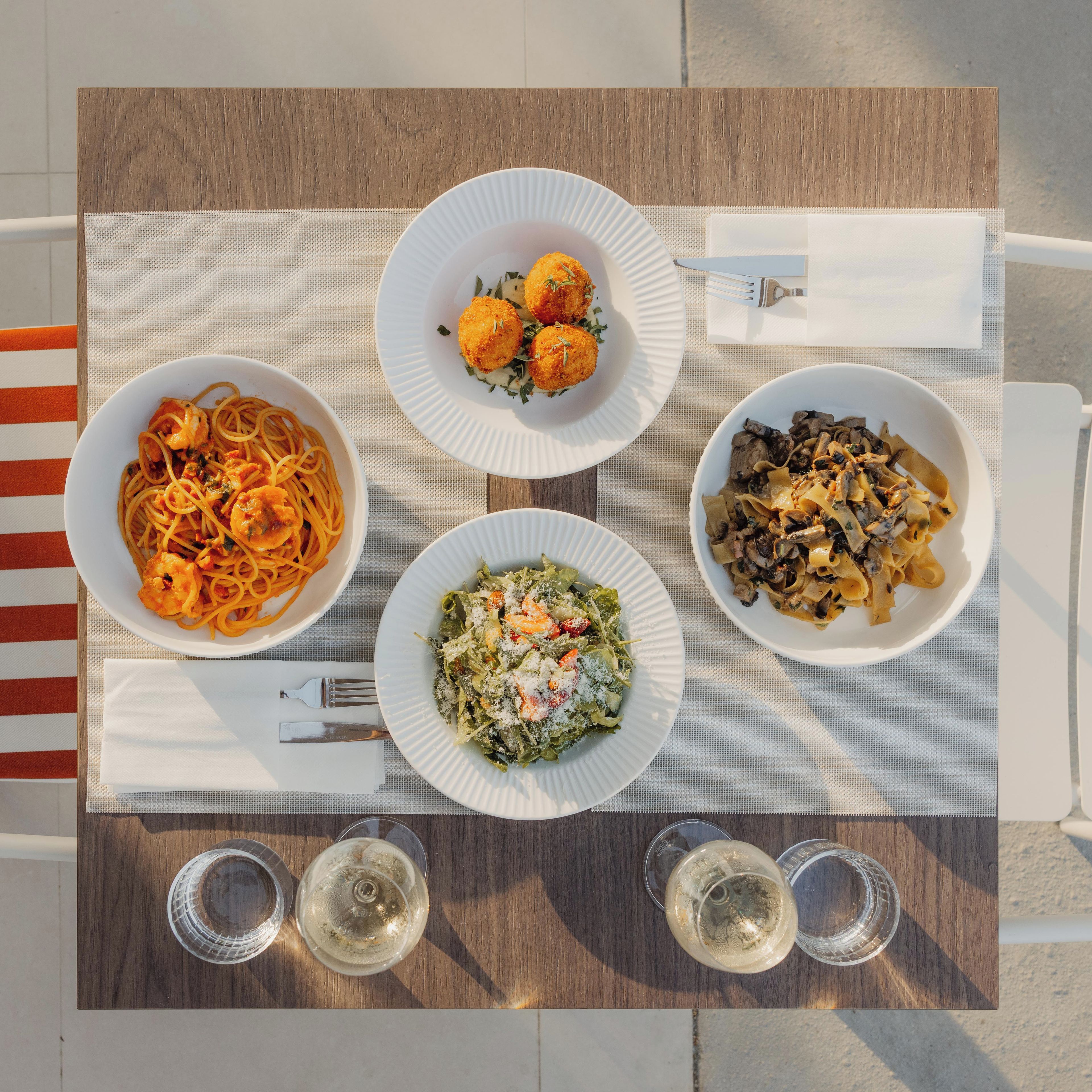 A table with four plates of pasta dishes, two empty glasses, and a striped napkin.