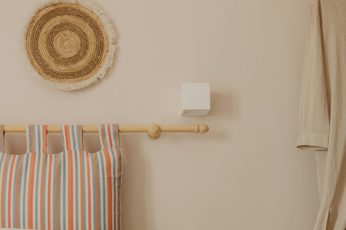 A bedroom with a bed, striped curtains, a wooden headboard, a round wooden side table with decorative items, and a wall-mounted towel rack.