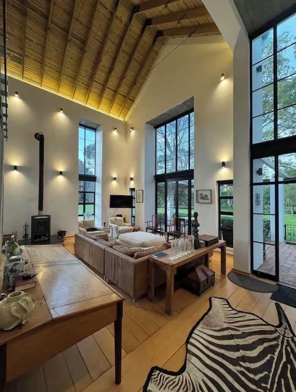 A spacious living room with a large sofa, a wooden dining table, and a fireplace. There are large windows providing natural light and views of the outdoors.