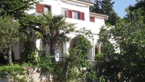 Ein weißes Gebäude mit roten Fensterläden, umgeben von viel Grün und Bäumen.
