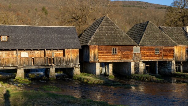 A rustic wooden building with a stone foundation, a small window, and a thatched roof, situated by a calm river. - Villa Vila Velebita - Three Bedroom Countryside Villa (Otocac)
