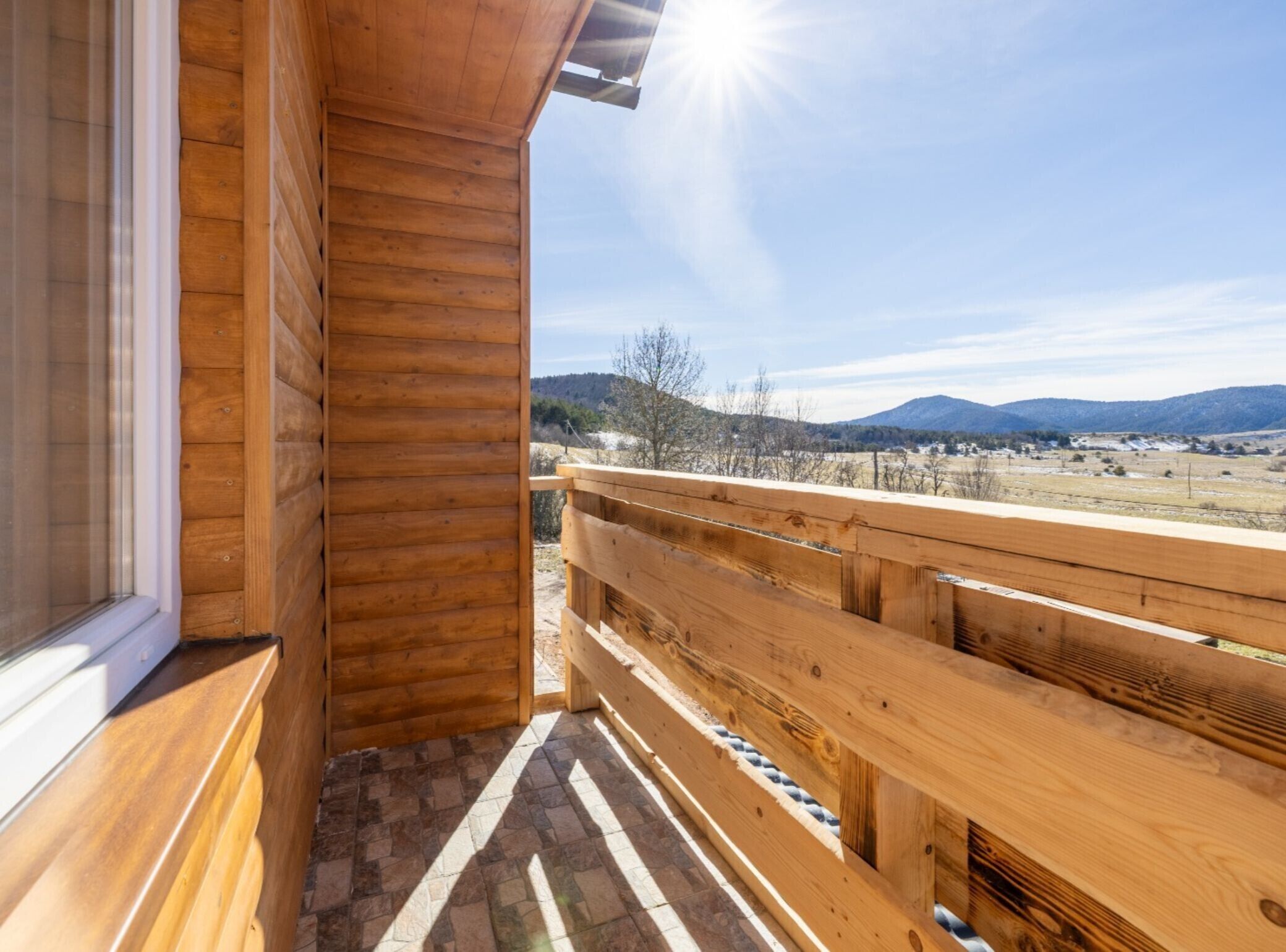 A wooden balcony with a stone-tiled floor, overlooking a rural landscape with mountains in the distance.