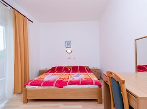 Een eenpersoonsbed met een rood-wit gestreepte deken, een houten nachtkastje, een bureau met een stoel en een raam met gordijnen.