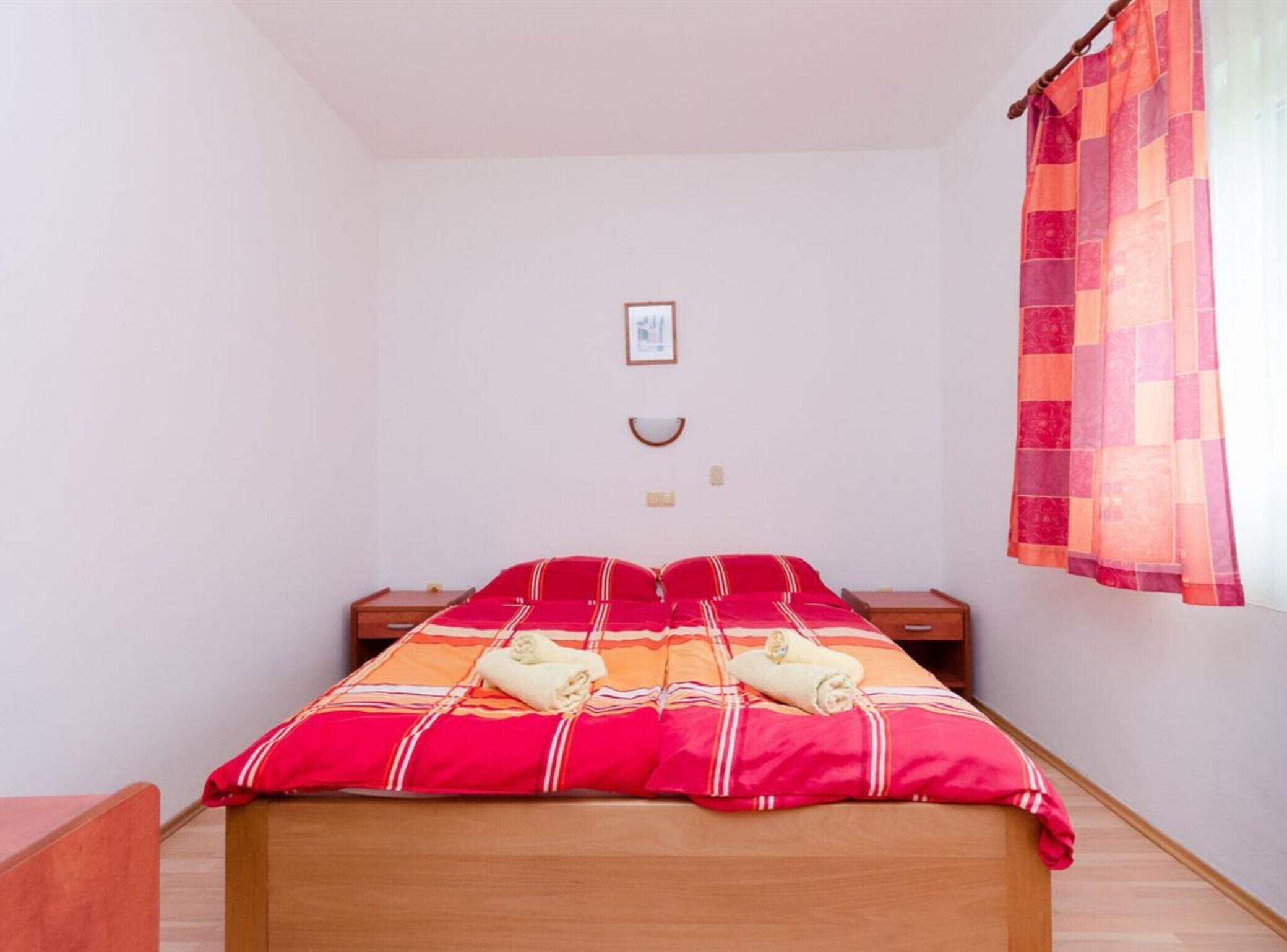 Una cama individual con colcha a rayas rojas y naranjas, dos mesitas de noche, un cuadro pequeño en la pared y una ventana con cortina a cuadros.