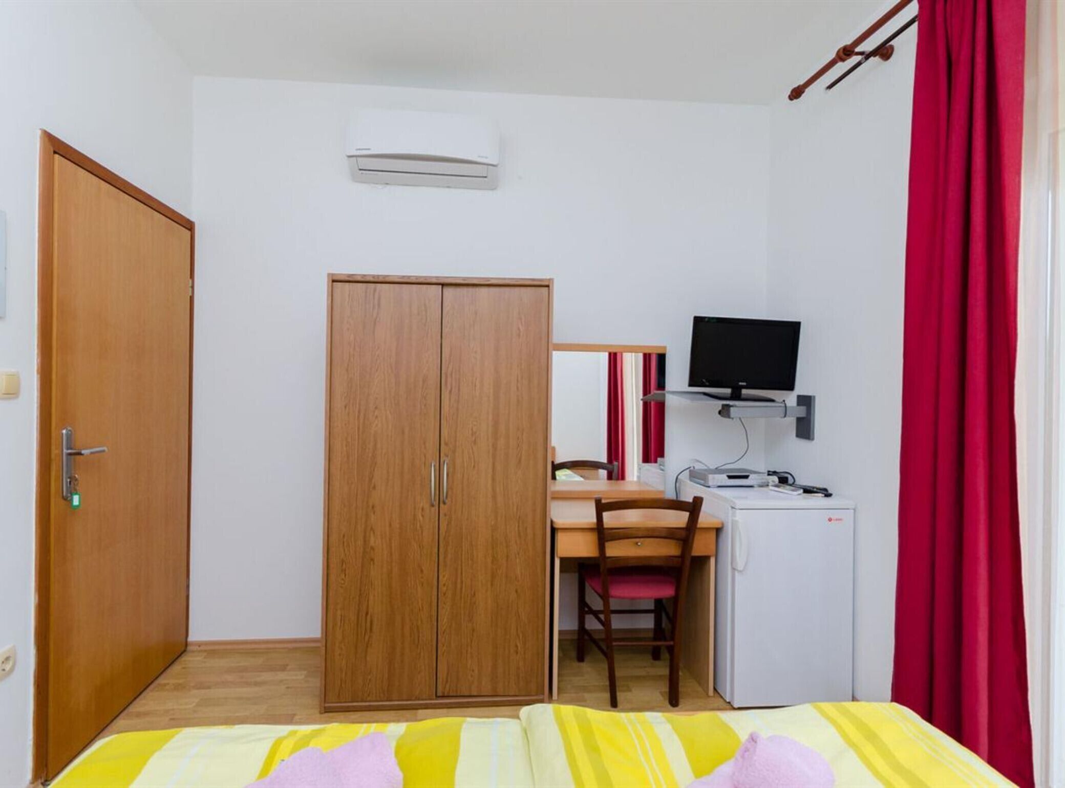 Une chambre de taille modeste, équipée d’une armoire, d’un bureau avec une chaise, d’une télévision et d’un petit réfrigérateur.