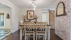 Uma sala de jantar com uma mesa e cadeiras brancas, um lustre pendente e uma parede de tijolos rústicos.