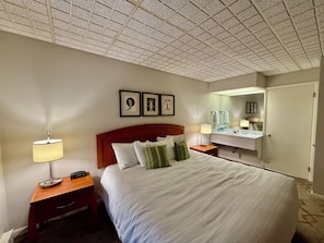 A hotel room with a large bed, a nightstand with a lamp, a wooden desk, a mirror, and framed artwork on the wall. - Lindsey Place (Lock Haven)